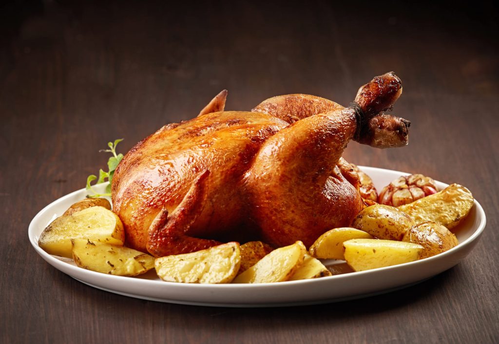 Pollo asado entero con papas al horno en un plato sobre mesa de madera.